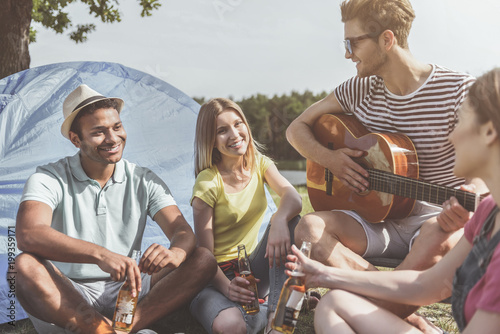 Lovely weekend. Delighted group of people sitting near tent with beer and guitar. They are laughing
