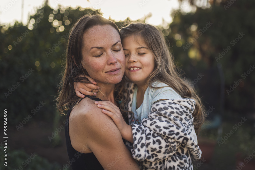 Mother and daughter with eyes closed embracing in yard during sunset Mother and daughter with eyes closed embracing in yard during sunset