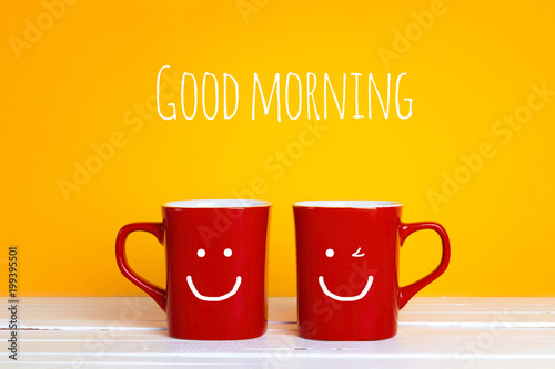 Two red coffee mugs with a smiling faces on a yellow background with the phrase Good morning.