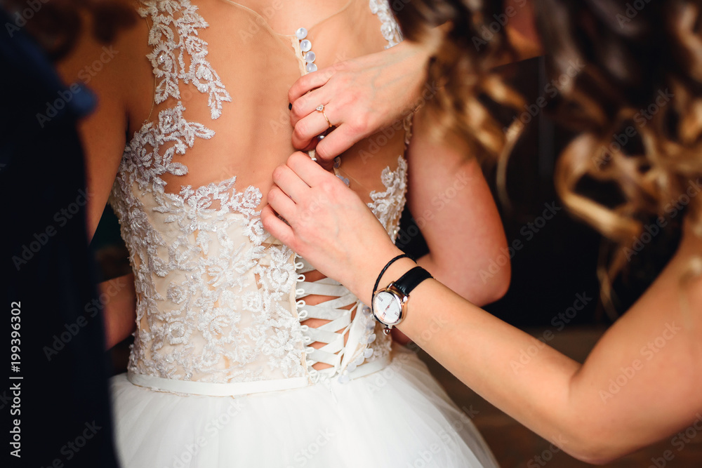 bridesmaid hands fastens buttons on the back of the bride on the wedding dress with beige corset close up
