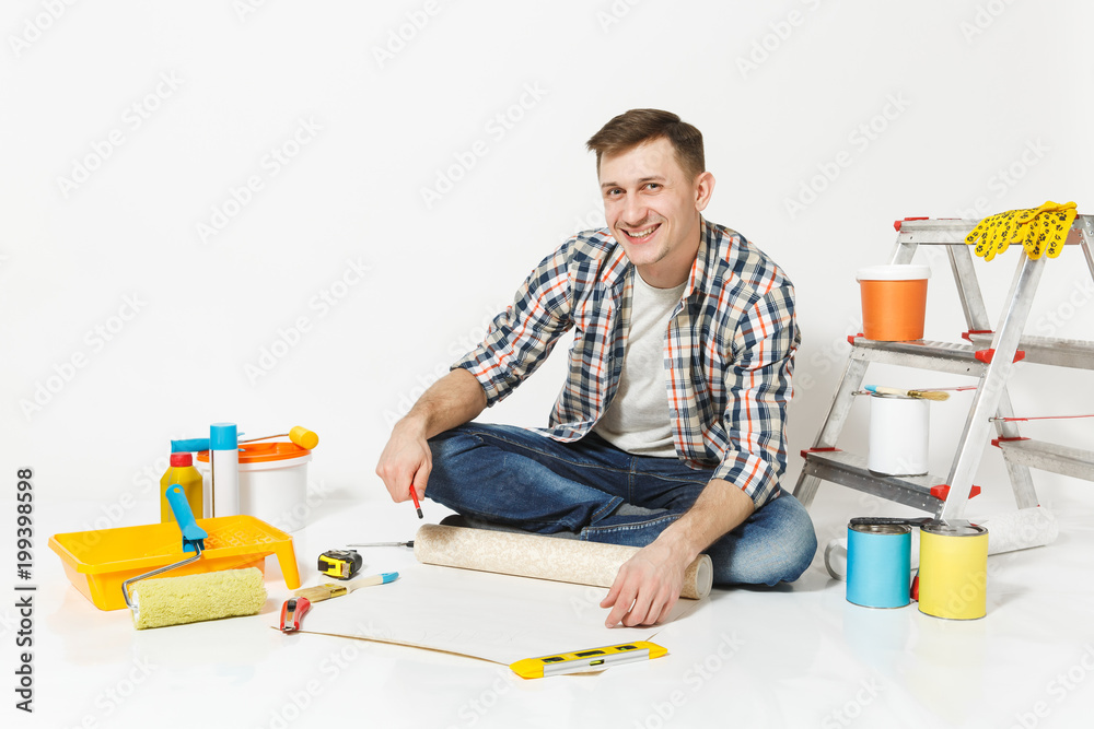 Young man sitting on floor with wallpaper roll, pencil, instruments for renovation apartment room isolated on white background. Wallpaper, gluing accessories, painting tools. Repair home concept. Young man sitting on floor with wallpaper roll, pencil, instruments for renovation apartment room isolated on white background. Wallpaper, gluing accessories, painting tools. Repair home concept.