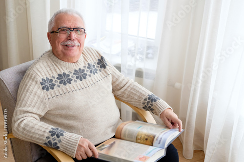 Senior man 75-75 years old sitting at home reading book in armchair