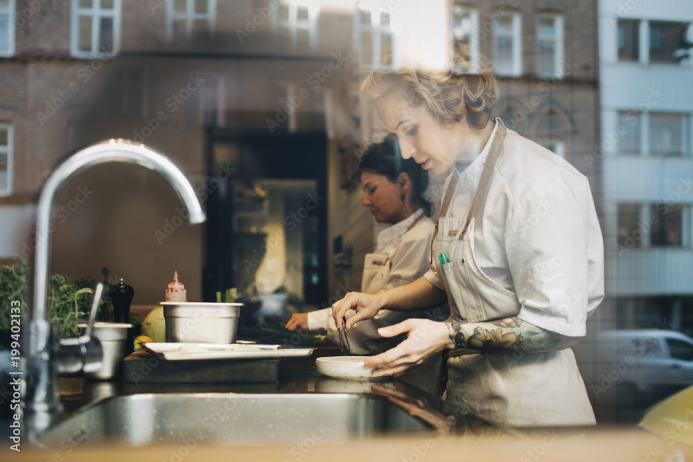 Side view of female chefs preparing food at counter in restaurant kitchen seen from window Side view of female chefs preparing food at counter in restaurant kitchen seen from window