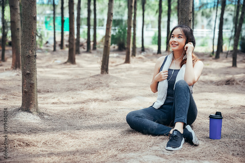 Asian girl is exercising, and drinking cool water and listening music at the weekend in a pine forest green and lush beautiful. Sport girl and lifestyle concept.