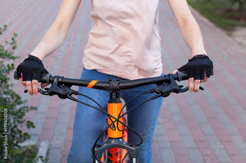 Fototapeta Naklejka Na Ścianę i Meble -  A girl in bicycle gloves holds her hand on the handlebars, clamping the brakes. A bicycle element on a street background. Bicycle steering wheel with speed selector, cables and brakes.