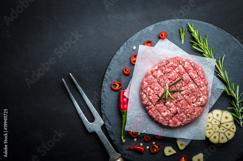 Organic raw ground beef patties for making homemade burgers with rosemary, species and carving fork on stone cooking background, top view, space for text