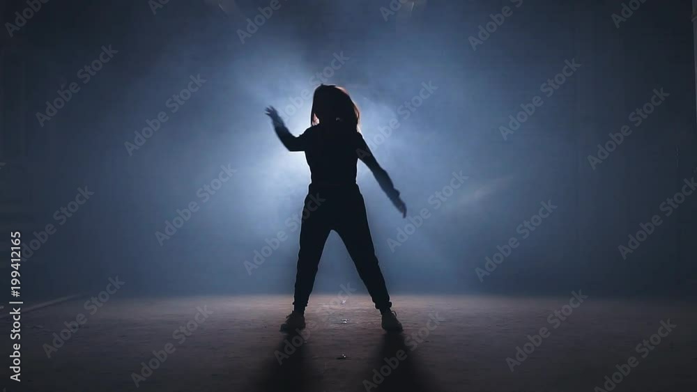 Smiling female in sporty clothers is demonstrating dance step. Trying a new dance step on smoky stage. Smiling female in sporty clothers is demonstrating dance step. Trying a new dance step on smoky stage.
