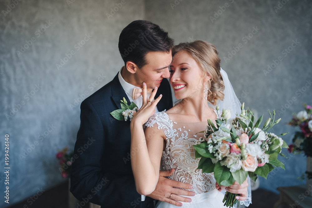 A stylish bridegroom cute embraces a bride in a lace dress. The newlyweds embrace in a gray studio with a beautiful bouquet. A stylish bridegroom cute embraces a bride in a lace dress. The newlyweds embrace in a gray studio with a beautiful bouquet.