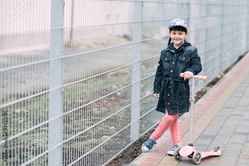 Cute little girl in jeans cardigan and on scooter on the background of the fence