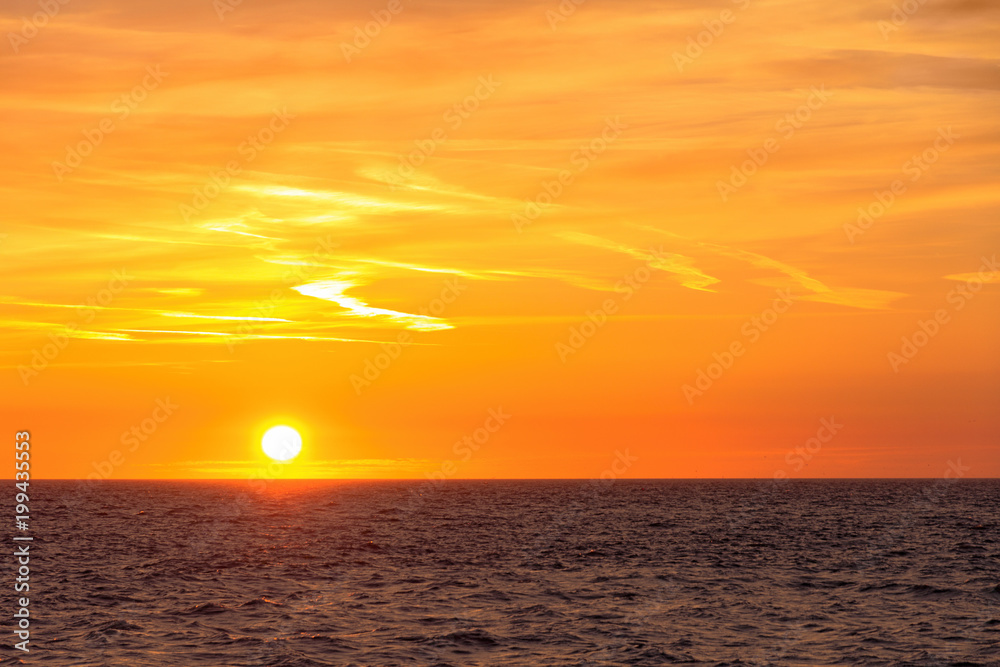 Beautiful round and bright sun setting against a vivid orange sky with high altitude tin clouds over the horizon of the North sea