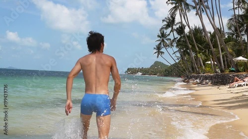 Back of view of young sporty man running on the beach at sunny dayand splashing sea waves on small droplets . slow motion. 1920x1080 photo