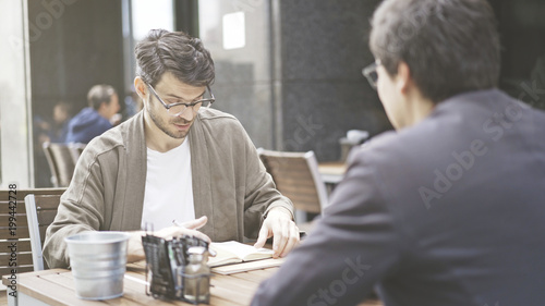 Two friends are having a conversation at table of the cafe outdoors. A man dressed in a jacket wearing eyeglasses looking into his papers in front of his friend