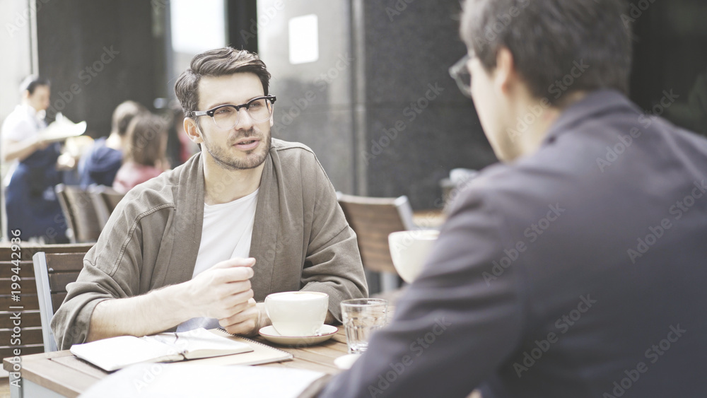 Two friends are having a conversation at table of the cafe outdoors. A man dressed in a jacket wearing eyeglasses is listening carefully to his friend talking Two friends are having a conversation at table of the cafe outdoors. A man dressed in a jacket wearing eyeglasses is listening carefully to his friend talking