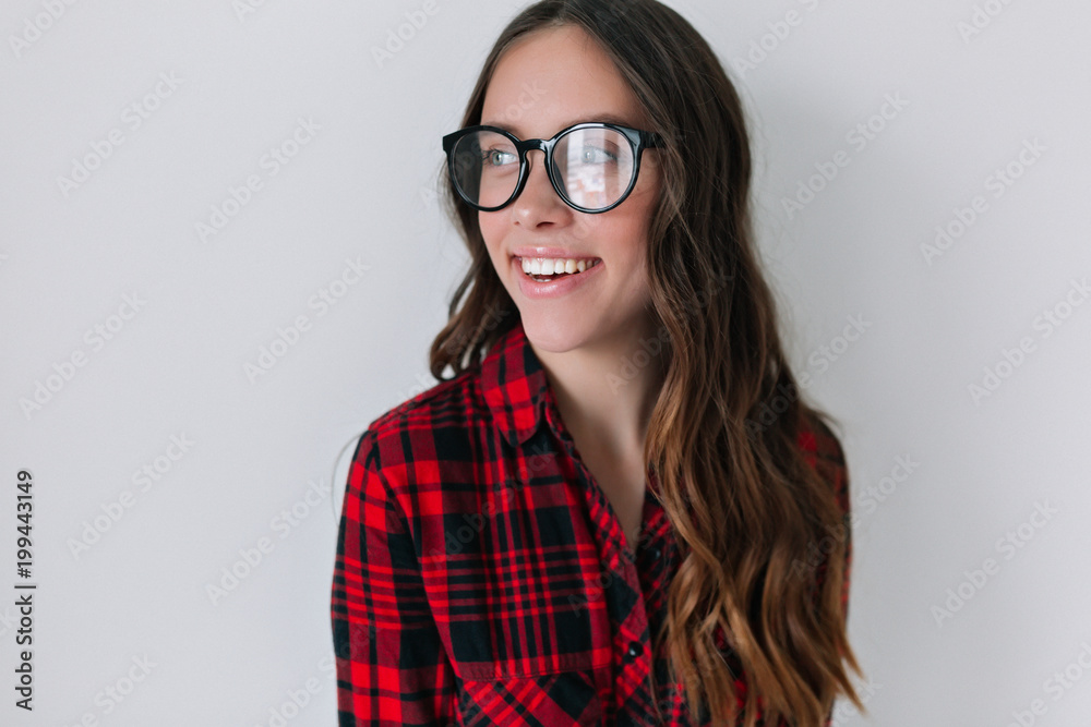 Indoor close-up portrait of charming pretty woman with nude make-up and long dark hair wears glasses and  checkered shirt smiles and looking at window against grey wall
