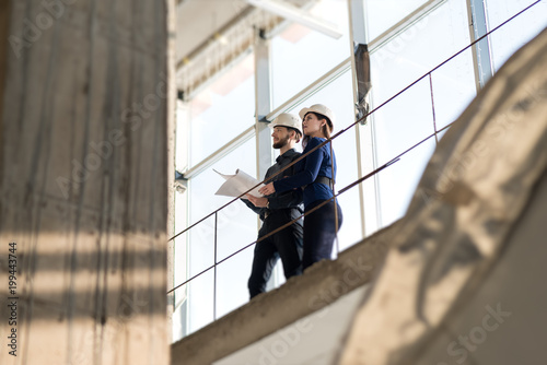 Two engineers, a man and a woman in construction helmets looking at a construction project. In white construction helmets standing on a construction bridge. Photographed from below.