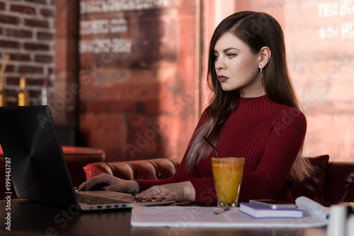 Beautiful business woman sitting with laptop in cafe. Prints on the keyboard. Red Turtleneck. A glass with juice with a straw. Notebook.
