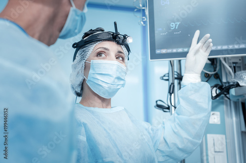 Heart beating rate. Positive nice pleasant surgeon holding her hand up and pointing at the monitor while checking patients heart beating rate