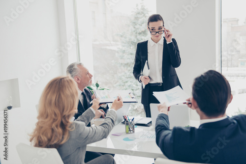 Beautiful  elegant  stylish  attractive woman in suit holding eyelet og glasses on her face  looking out spectacles to her colleagues who giving clipboard with diagrams  having disagreement