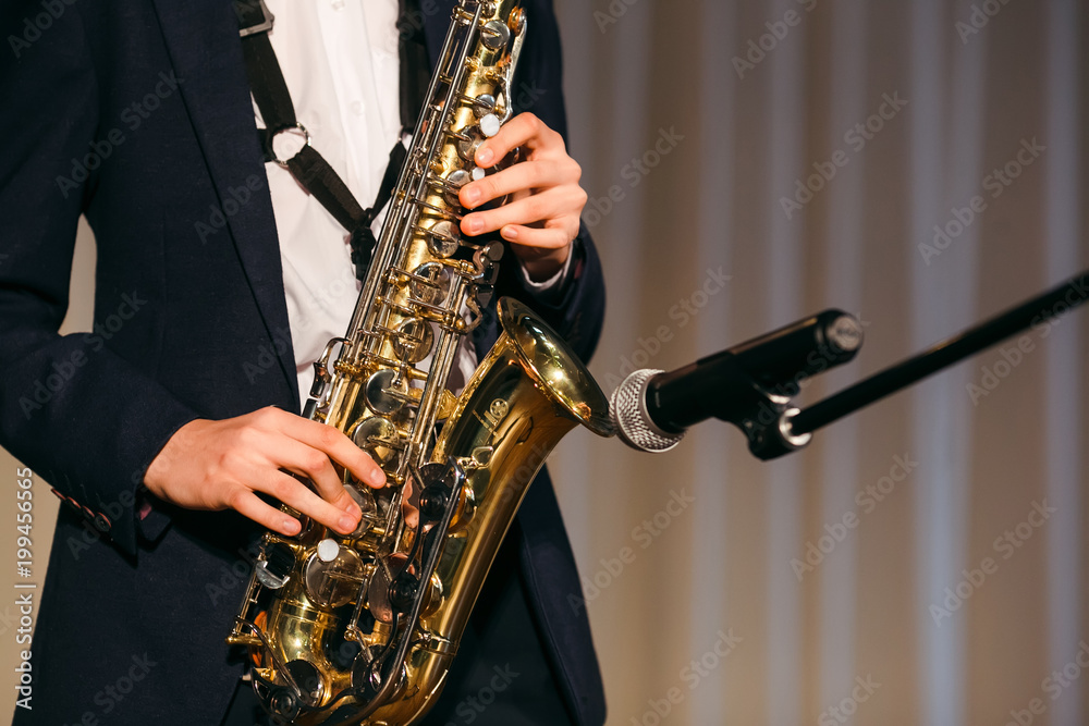 Obraz premium Young boy hands in black jacket and white shirt playing saxophone at the microphone on stage