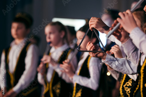  Little girl hands is playing on a musical triangle at stage close-up. Children orchestra in Tatar national clothes and skullcaps plays on the national instrument Kurai