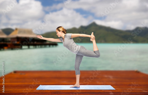 fitness, sport, people and healthy lifestyle concept - woman making yoga in lord of the dance pose on wooden pier over island beach and bungalow background