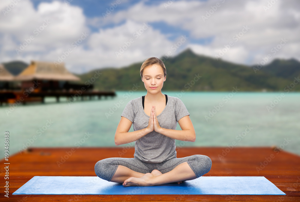 fitness, meditation and healthy lifestyle concept - happy woman making yoga lotus pose on wooden pier over island beach and bungalow background