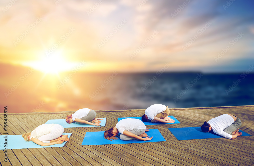 fitness and healthy lifestyle concept - group of people doing yoga child pose outdoors on wooden pier over sea background fitness and healthy lifestyle concept - group of people doing yoga child pose outdoors on wooden pier over sea background