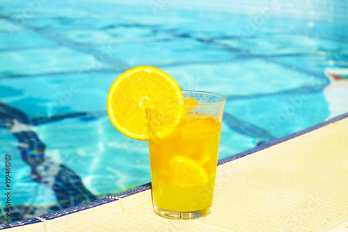 Close up of screwdriver cocktail alcohol drink with orange juice, slices and ice standing near the pool. Refreshing iced lemonade beverage in glass by the poolside. Sun glares. Background, copy space.