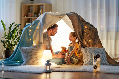 family, hygge and people concept - happy father with teddy bear toy and little daughter playing in kids tent at night at home photo