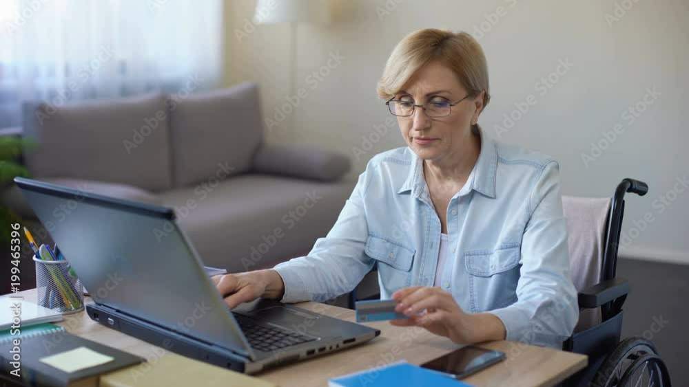 Nice senior woman in wheelchair typing card number on laptop, paying bills Nice senior woman in wheelchair typing card number on laptop, paying bills