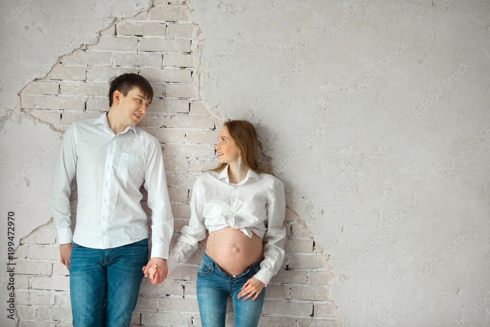 beautiful husband and pregnant wife in jeans and white shirt standing near white wall