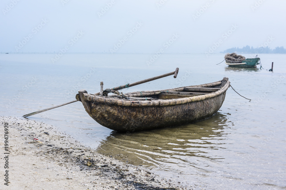 Bamboo and wood traditional fishing boats tied up on the beach at Lap An Lagoon, Vietnam, with a large oyster farm in the mist behind.