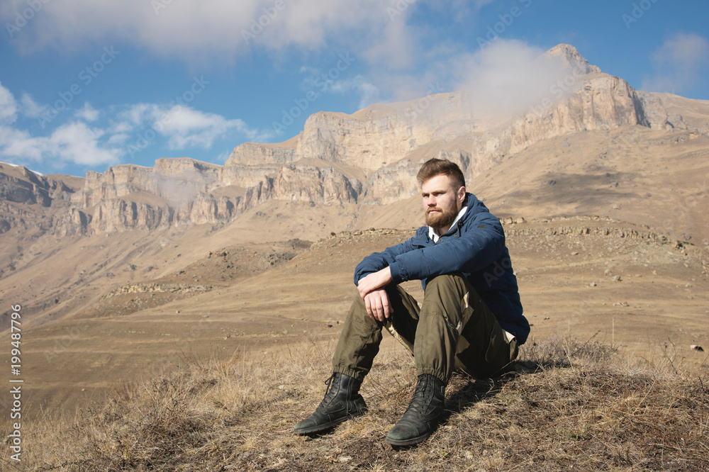 A portrait of a stylish bearded hipster sitting on a rock against the backdrop of epic rocks and contemplating into the distance thinking about life. The concept of tranquility and solitude with A portrait of a stylish bearded hipster sitting on a rock against the backdrop of epic rocks and contemplating into the distance thinking about life. The concept of tranquility and solitude with
