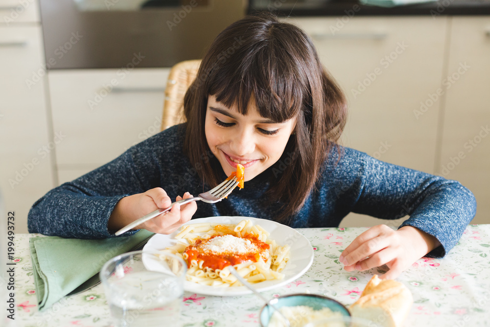 Cute and happy little girl eating pasta with tomato sauce and powdered cheese in the kitchen at home Cute and happy little girl eating pasta with tomato sauce and powdered cheese in the kitchen at home