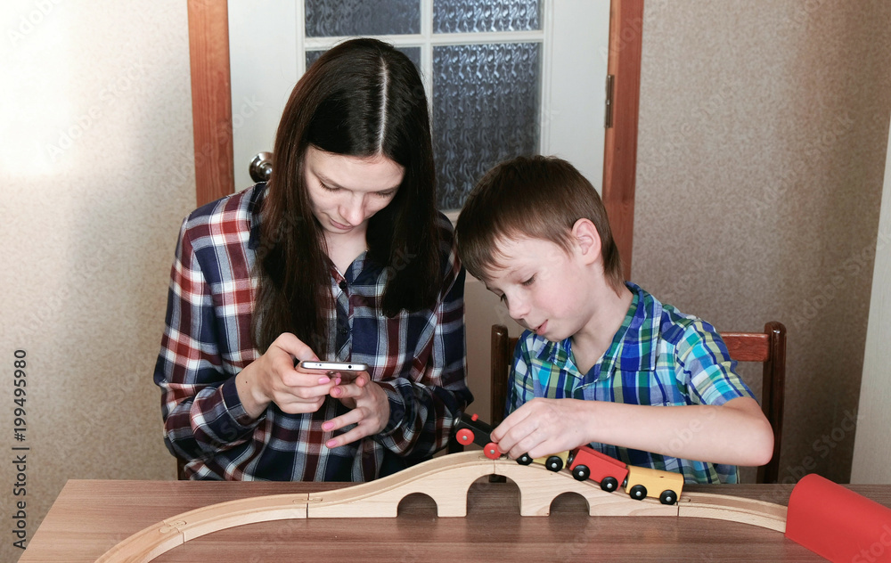 Fototapeta premium Playing together. Mom looks at phone and son is playing a wooden railway with train, wagons and tunnel sitting at the table.