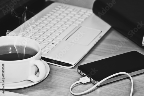 Black and white photo of a cup of hot tea on the table next to the laptop and phone