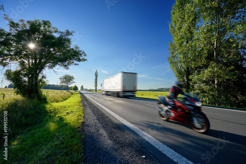 Obraz na płótnie Motion blurred motorcycle and a truck driving on asphalt road with the sun shining through the trees in a rural landscape w salonie