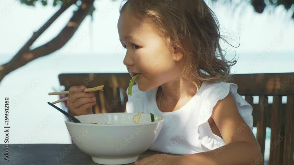 Child girl eats noodles soup via chopsticks at the open air beach cafe Child girl eats noodles soup via chopsticks at the open air beach cafe