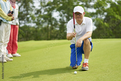 Asian golfer crouching in golf course aiming and preparing for putting