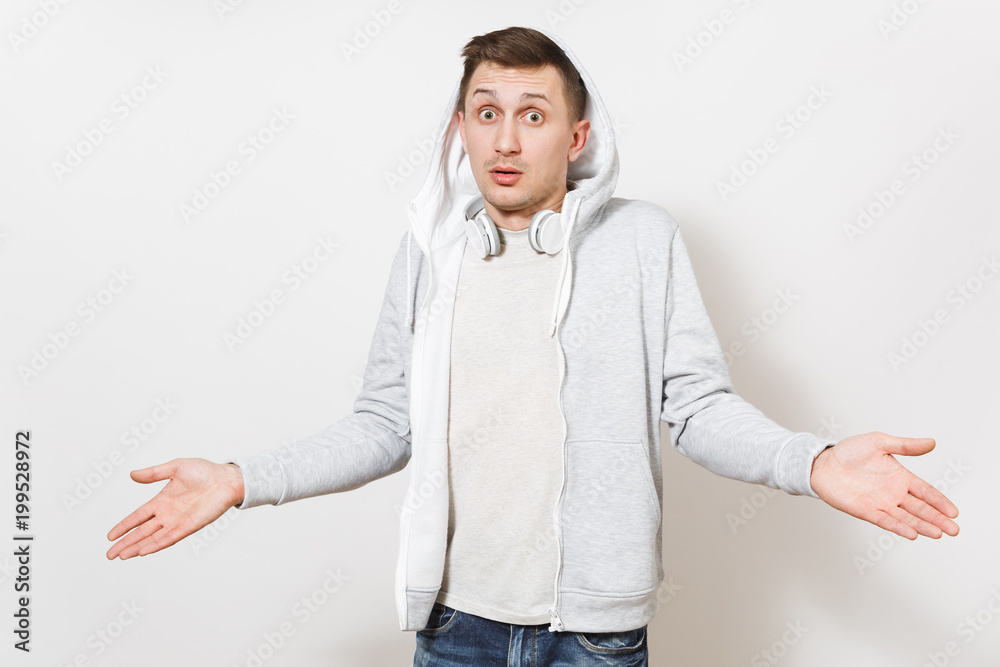 Young handsome upset man student in t-shirt and light sweatshirt with hood with headphones with puffed face spreads hands in different directions in studio on white background. Concept of emotions Young handsome upset man student in t-shirt and light sweatshirt with hood with headphones with puffed face spreads hands in different directions in studio on white background. Concept of emotions