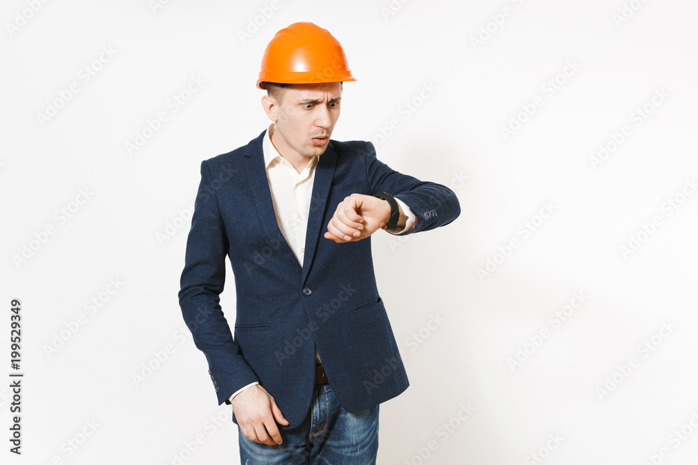 Young irritated dissatisfied businessman in dark suit, protective construction orange helmet looking on clock on hand isolated on white background. Time is running out. Male worker for advertisement. Young irritated dissatisfied businessman in dark suit, protective construction orange helmet looking on clock on hand isolated on white background. Time is running out. Male worker for advertisement.