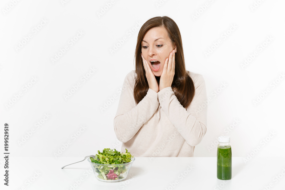 Shocked woman sitting at table with green detox smoothies, salad in glass bowl isolated on white background. Proper nutrition, vegetarian food, healthy lifestyle, dieting concept. Area with copy space Shocked woman sitting at table with green detox smoothies, salad in glass bowl isolated on white background. Proper nutrition, vegetarian food, healthy lifestyle, dieting concept. Area with copy space