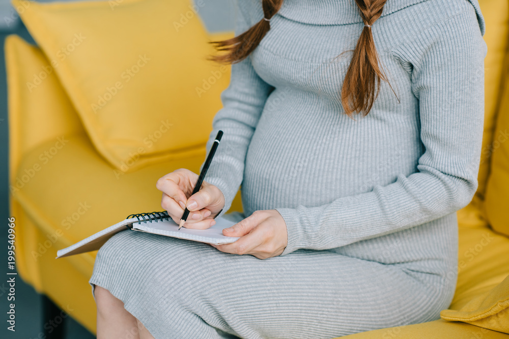 cropped image of pregnant woman writing something to notebook on sofa