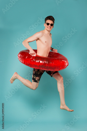The summer, when it arrives, is the more gratefully received! Attractive young boy with naked torso jumping with rubber ring over isolated blue background.