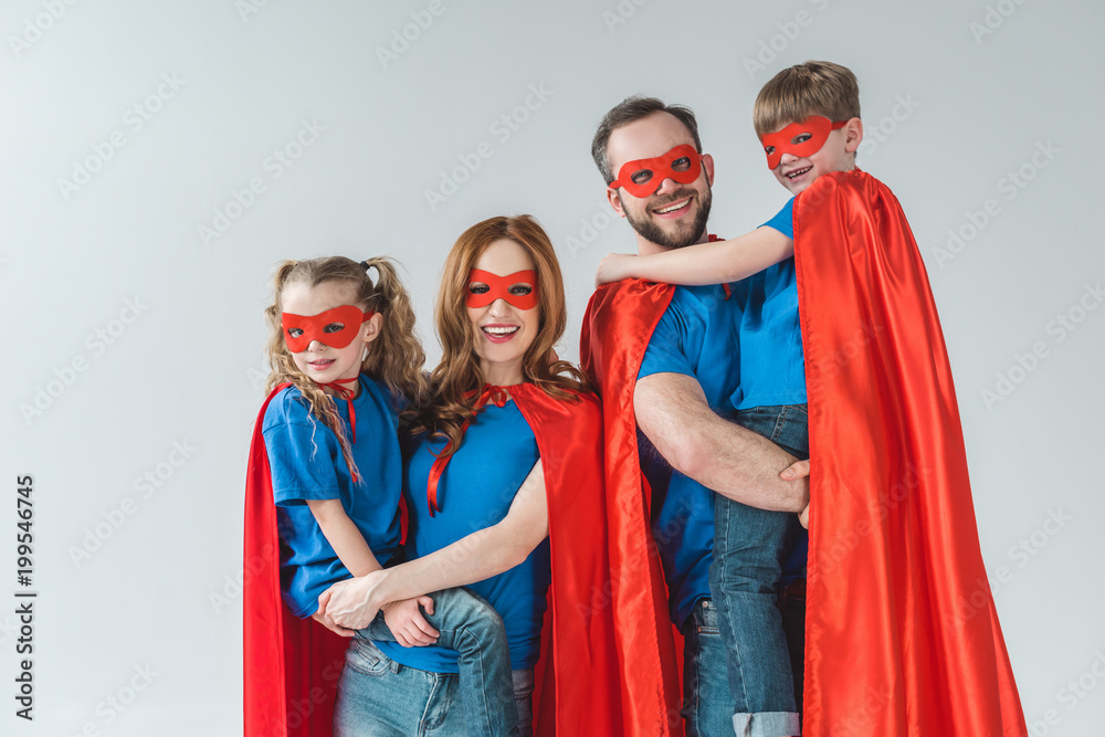 cheerful family of superheroes in masks and cloaks smiling at camera on grey
