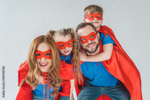 super family in masks and cloaks having fun together and smiling at camera isolated on grey