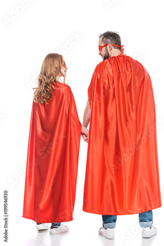 rear view of couple in masks and cloaks holding hands and looking at each other isolated on white