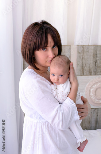 Obraz na płótnie Young mother is holding her little baby. Woman hugs her newborn child. Bit sad look. White light interior w salonie