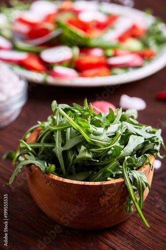 Fresh ruccola in a wooden bowl on wooden table over vegetable background, shallow depth of field.
