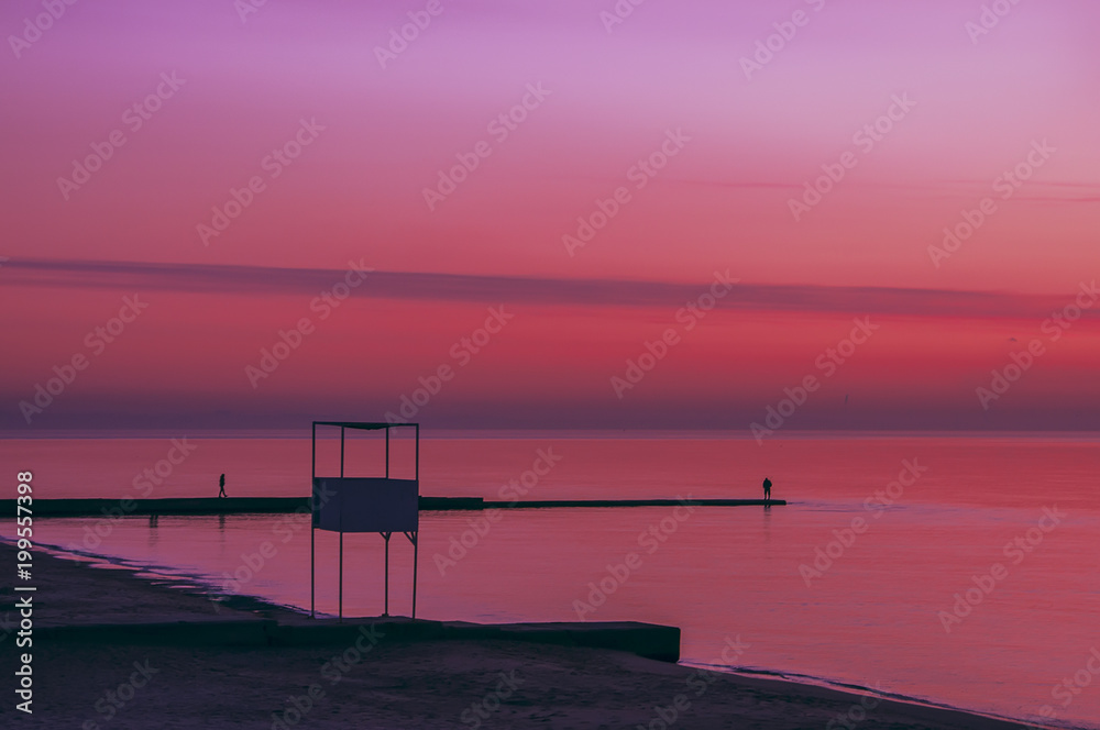 Incredibly colorful dawn on the sea. The dark silhouette of the tower, the sea line, the man on the pier.
